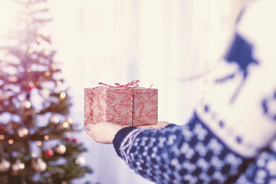 A hand in a patterned sweater holding out a small wrapped gift in front of a lit Christmas tree