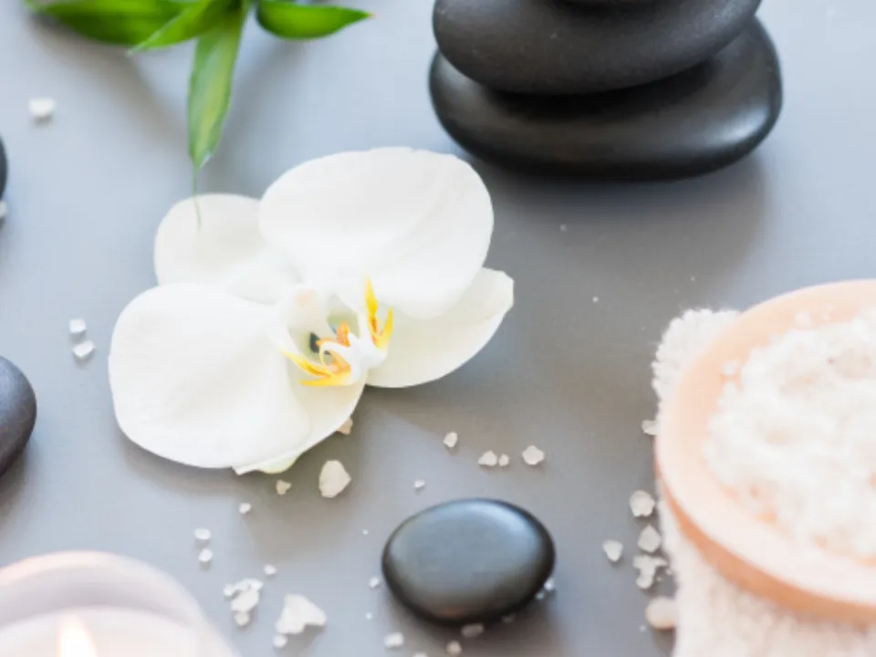 Black massage stones, a white orchid, a lit candle and a bowl of bath salt on a folded towel